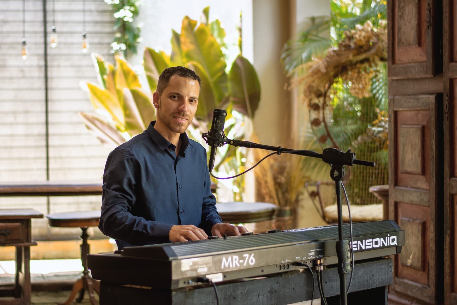 Jonathan Lage de camisa azul tocando teclado Ensoniq MR-76 em ambiente com plantas e microfone