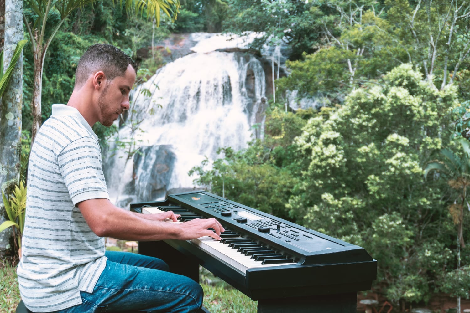 Jonathan Lage de camisa listrada tocando teclado ao ar livre diante de cachoeira em meio à mata